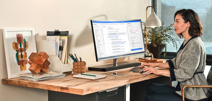 A dental clinic administrator sits nearby his work desk watching her computer screen while sitting in her office chair. On the table are a notebook, a phone, and a lamb.