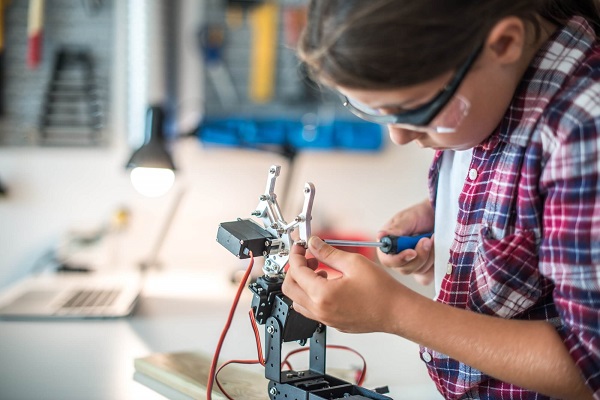 A little school kid is wearing a protection eye glass and doing some wiring work on a robot, with screw driver on her hand.