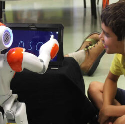 Two little kids are curiously watching a robot with white and orange color combo writing something on a tablet.