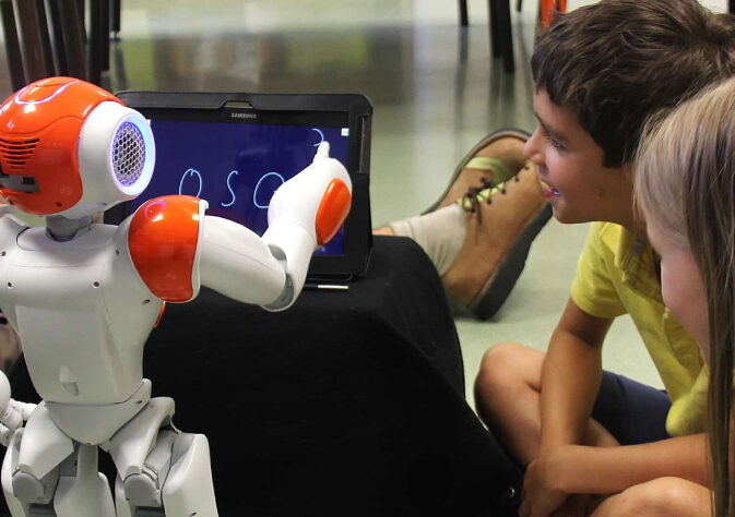 Two little kids are curiously watching a robot with white and orange color combo writing something on a tablet.