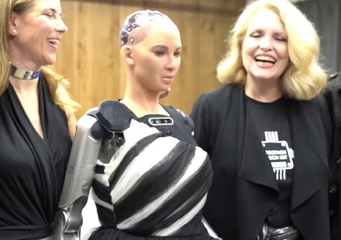 Two smiling women with a robot standing in between them